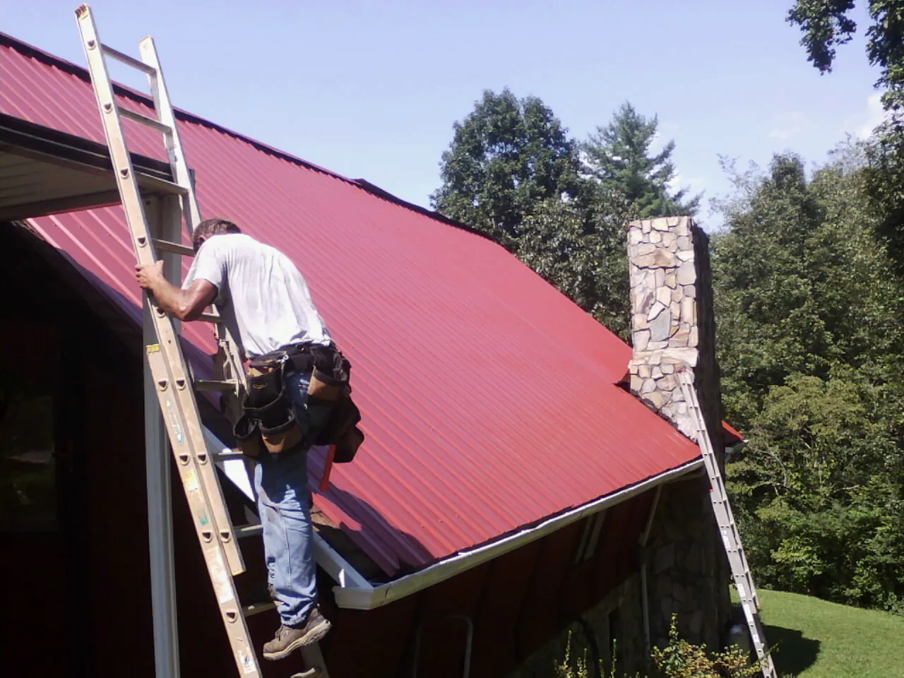 Roofer on a ladder beside a red metal roof and stone chimney — Soffit & Fascia Repair by Roofing Brothers in Malmo